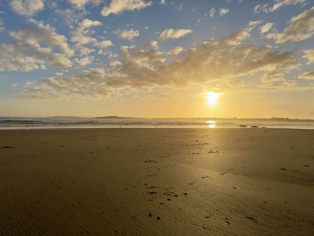 Wide sandy beach at sunset with scattered clouds, calm waves, and the sun low on the horizon.