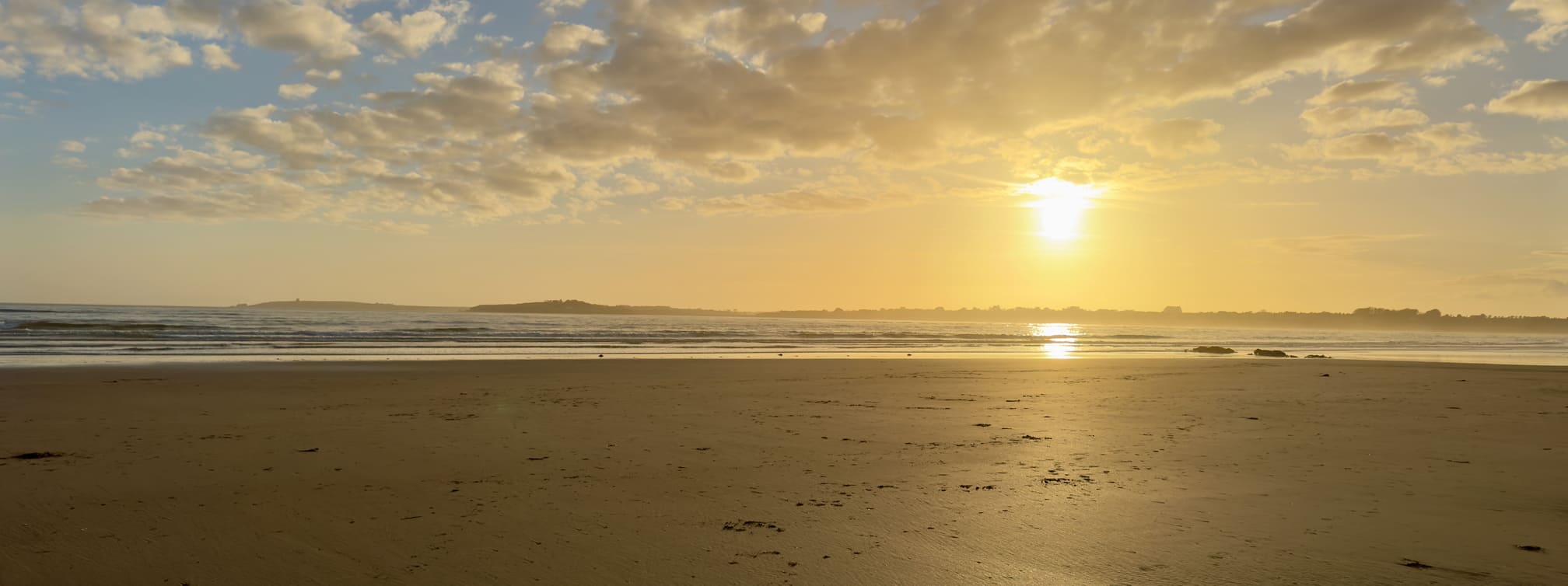A beach scene at sunset, with a clear sky and scattered clouds reflecting sunlight over the ocean, creating a warm glow on the sand.