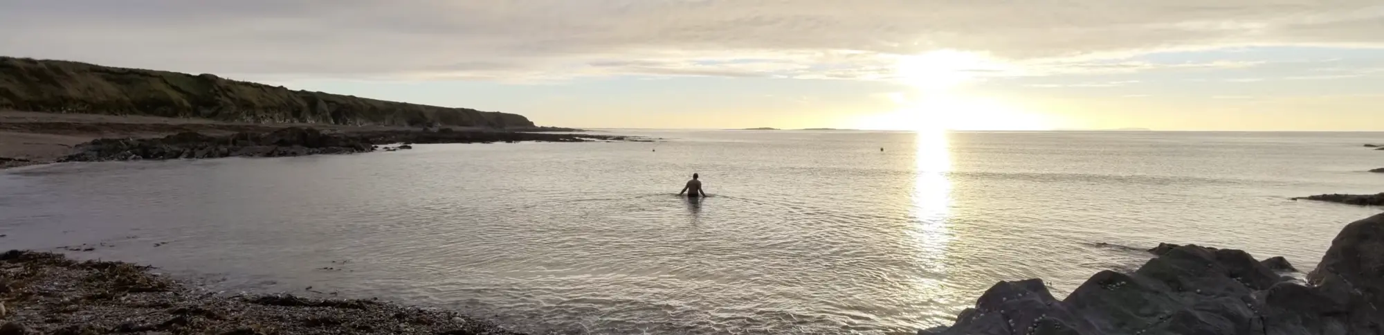 Kenn wades in a calm sea near rocky shorelines at sunrise, with the horizon stretching across the image.