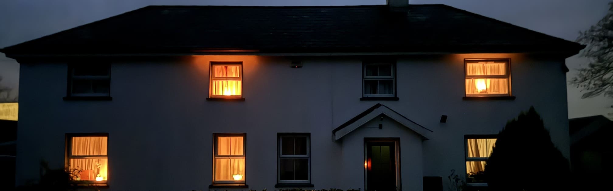 A two-story house at night with warm lights glowing from the windows, illuminating the dark exterior.