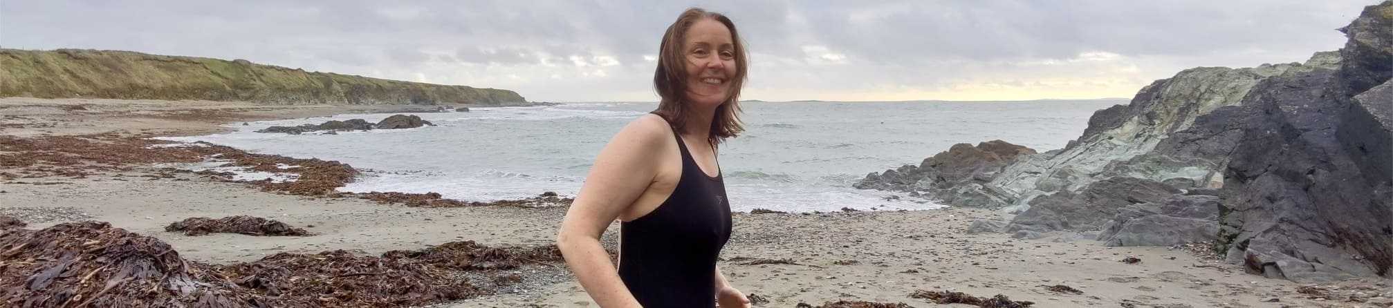 Helen in a black swimsuit walks along a rocky beach with seaweed, under a cloudy sky.