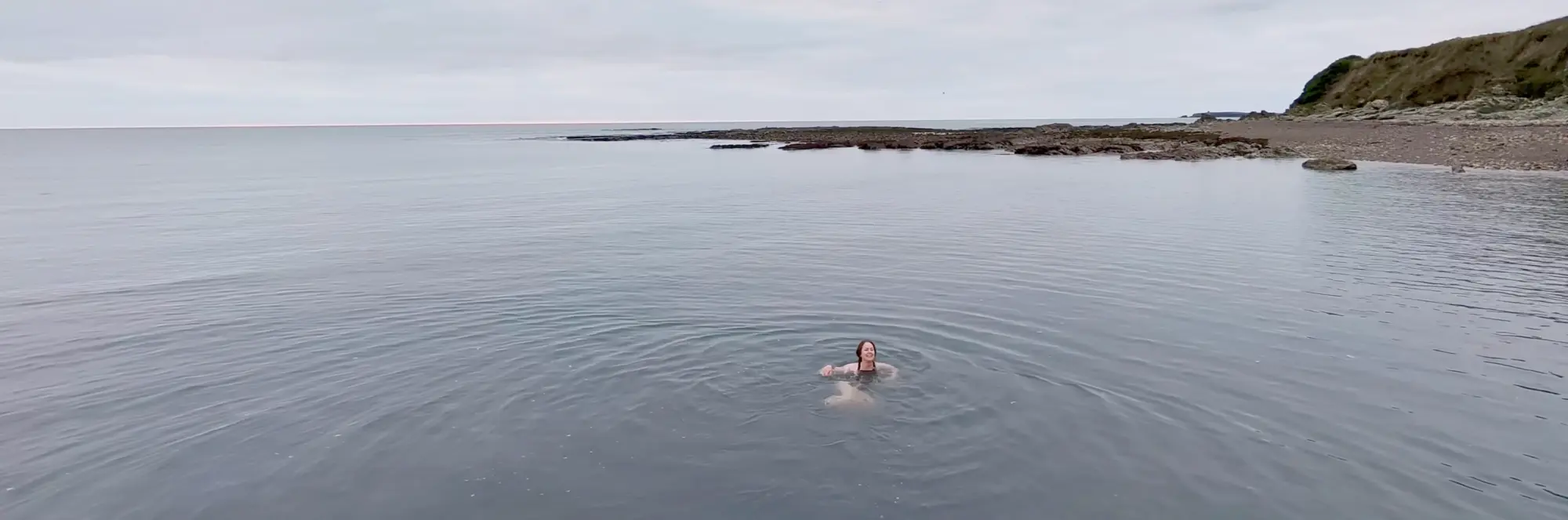 Helen swimming alone in a large, calm body of water near a rocky shoreline under an overcast sky.