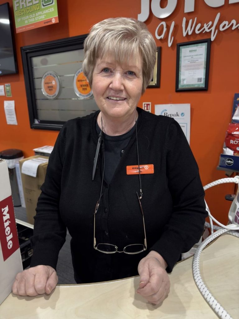 Breda in a black sweater is standing behind a counter, with a pair of glasses hanging around her neck. She is in a store with an orange sign in the background.