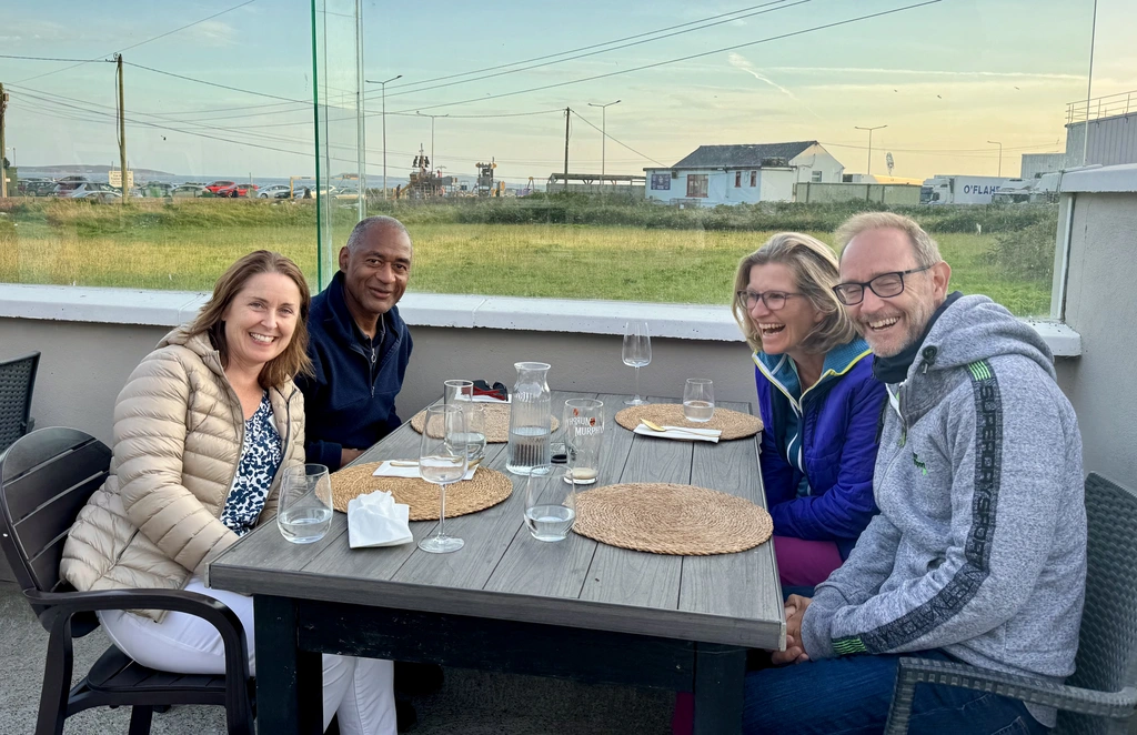 Helen, Kenn, Sabine and Peter sit around an outdoor table at The Ground Restaurant in Kilmore Quay with empty glasses and plates, smiling at the camera. A grassy field and buildings are visible in the background.