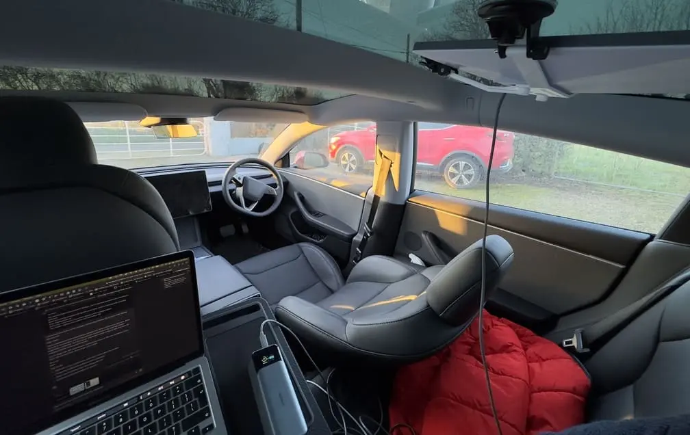 Interior of a my Tesla with a reclined front seat, a laptop in the back seat, and cables attached. Our MG ZS EV is visible through the window.