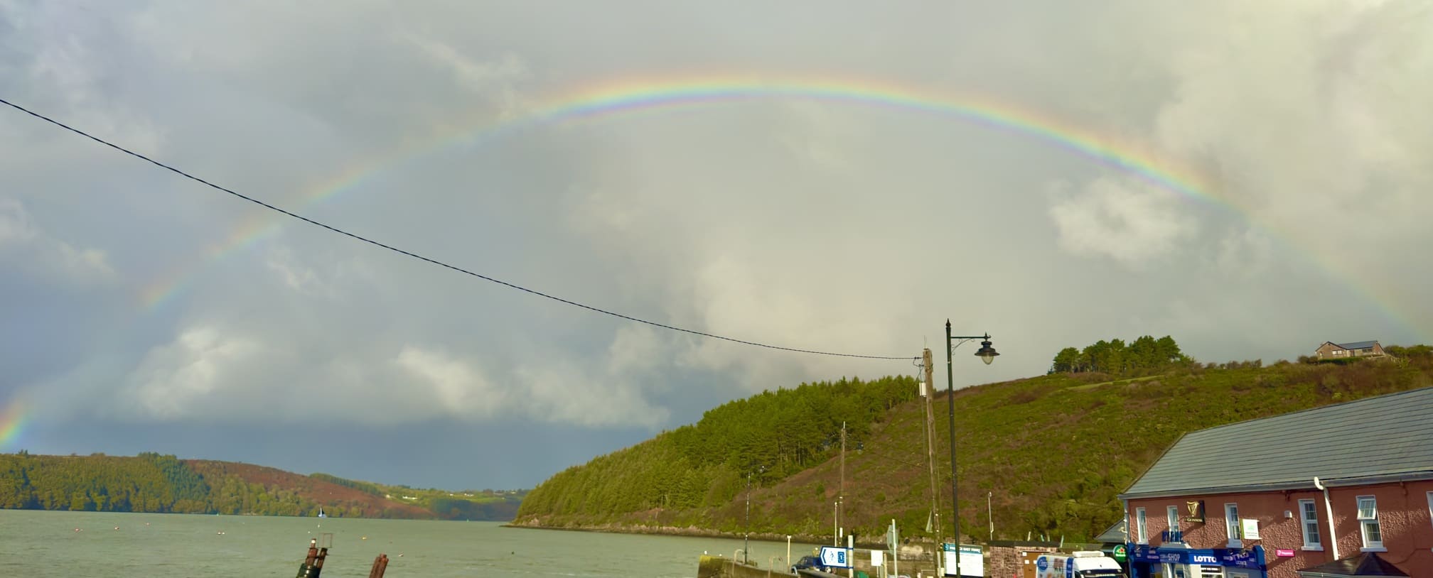 A rainbow arcs over a river, with lush green hills in the background and a few buildings near the water's edge.