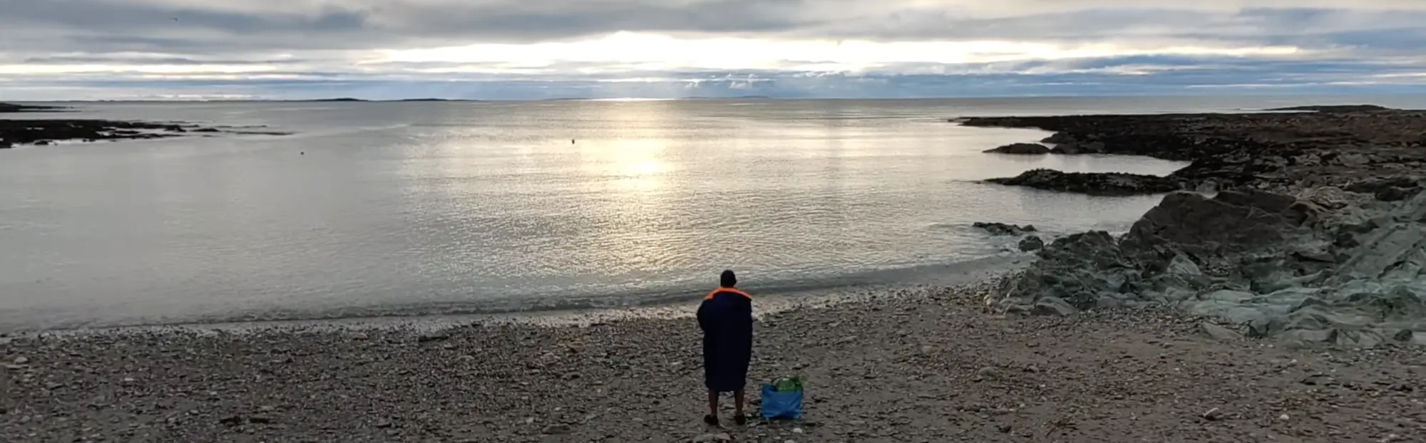Kenn stands on a rocky beach, looking out at a calm sea under a cloudy sky.