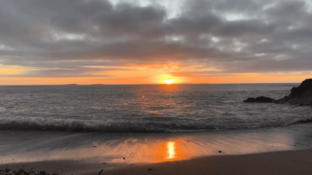 A sunrise over the Irish Sea with light reflecting on the wet sand, clouds partially covering the sky.