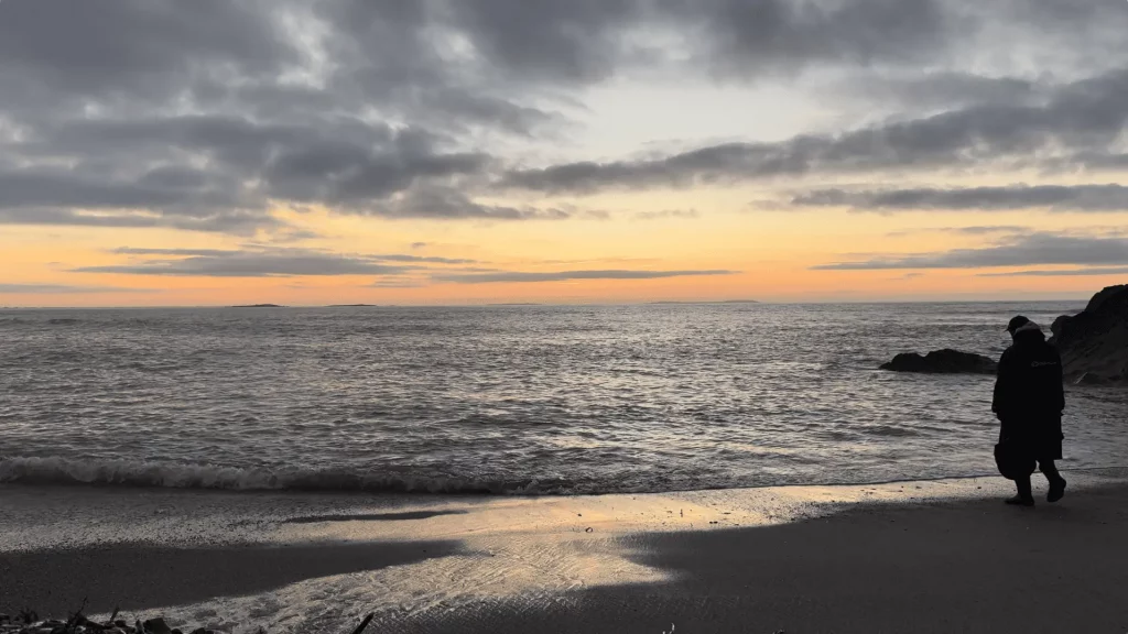 An overcast sky at sunrise casts reflected light on the ocean waves and wet sand. Kenn stands on the shore near rocks, facing the water.