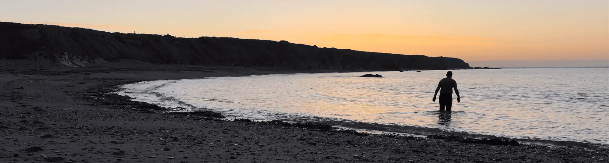 Kenn stands in shallow water near the shore at sunset, with cliffs and a calm sea in the background.