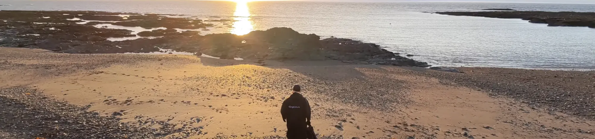 Kenn stands on a rocky beach, facing a calm sea with the sun setting on the horizon.