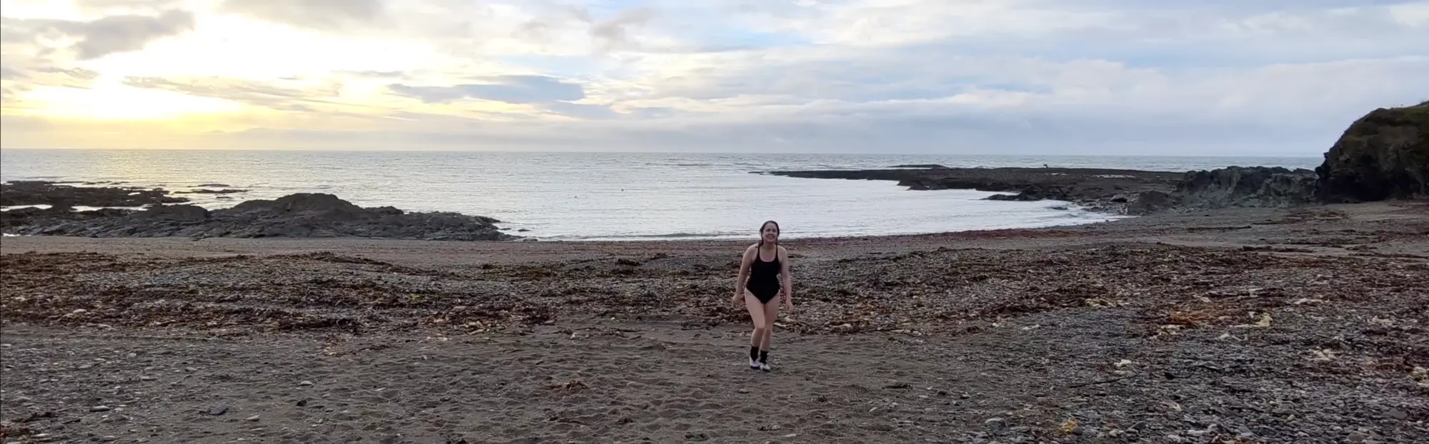 Helen in athletic wear walking on a rocky beach toward the camera, with the ocean and cloudy sky in the background.