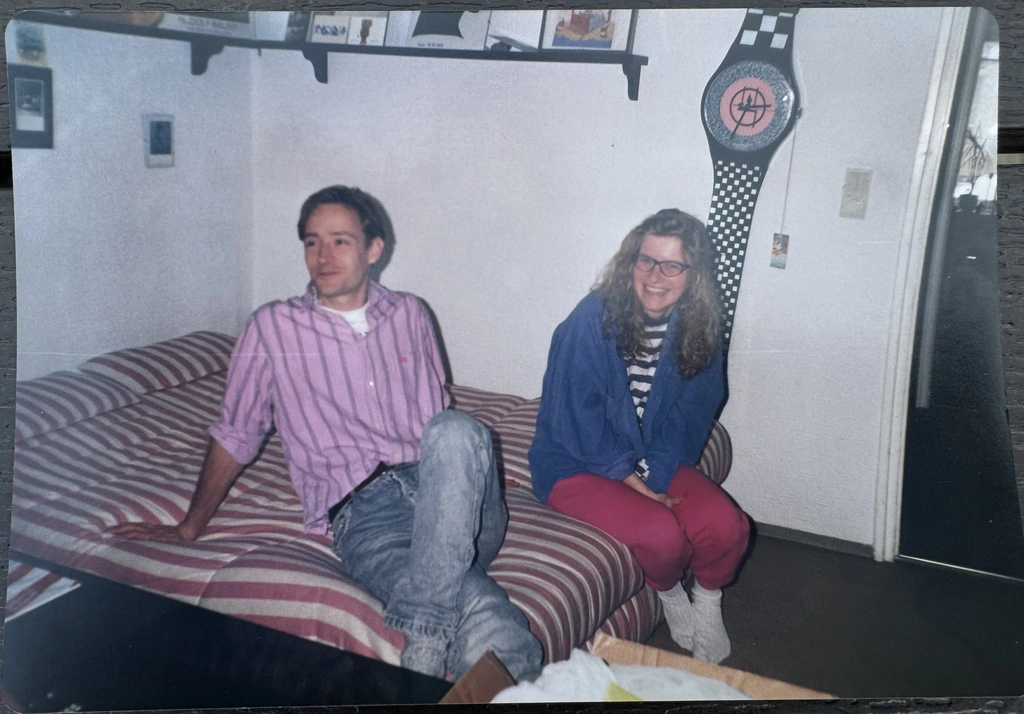 Two people sit on a striped couch in a room; the man wears a striped shirt and jeans, the woman wears glasses, a blue jacket, and red pants. A large decorative belt hangs on the wall.