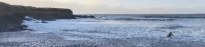 A person swimming in the ocean near a rocky shore with waves crashing and a cloudy sky in the background.
