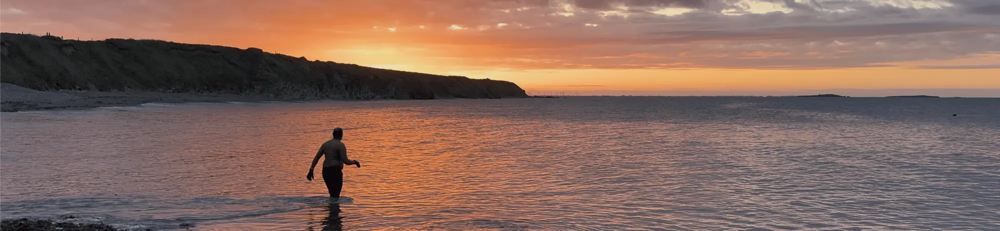 A person stands in shallow water at the beach during sunset, with a rocky coastline in the distance.